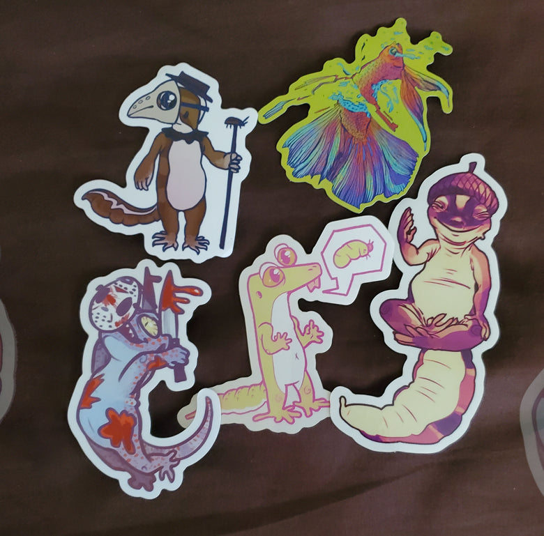 Five stickers, starting from top-left moving clockwise- AFT gecko in bird black plague mask with cockroach walking stick. Next is a red-blue betta fish torn in half, crying, the background is neon yellowish green. Next is AFT gecko wearing acorn shell as a hat and posing in god-like yoga. Next is pale yellow silly leopard gecko with speech bubble asking for waxworm. Next is a murderous tokay gecko with bloody knives, wearing jason the killer mask.