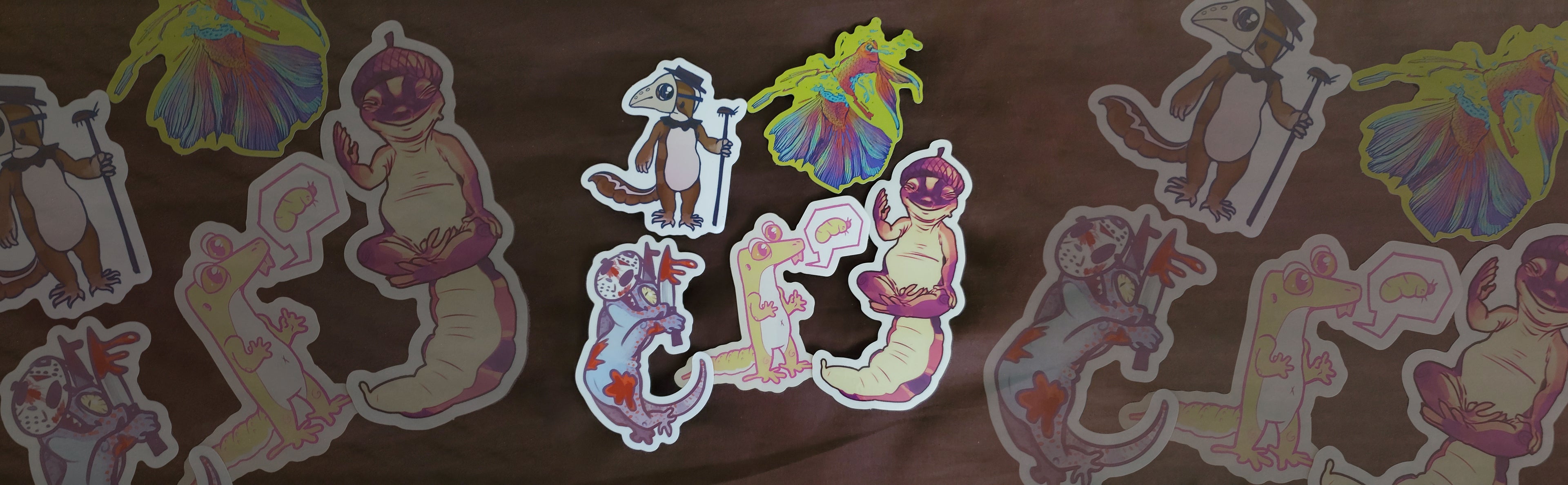A long banner version of a photo. Five stickers, starting from top-left moving clockwise- AFT gecko in bird black plague mask with cockroach walking stick. Next is a red-blue betta fish torn in half, crying, the background is neon yellowish green. Next is AFT gecko wearing acorn shell as a hat and posing in god-like yoga. Next is pale yellow silly leopard gecko with speech bubble asking for waxworm. Next is a murderous tokay gecko with bloody knives, wearing jason the killer mask.